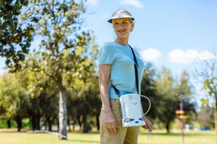 A woman walks in the park on a sunny day wearing her portable oxygen concentrator on a strap across her chest