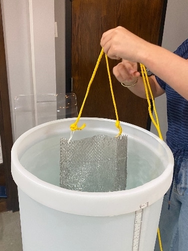 A mesh basket with hearing aids are lowered into a tub of water to test water resistance ratings
