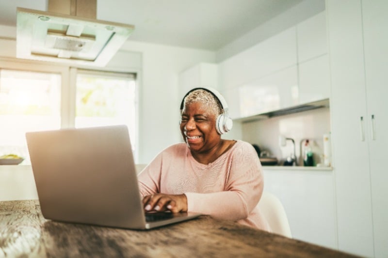 Woman smiling while wearing headphones and using her laptop at her kitchen table.