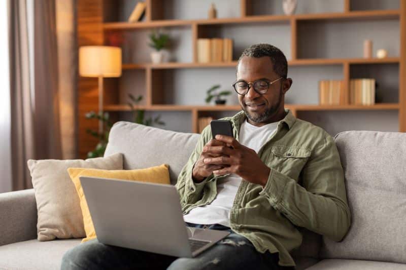 Adult man wearing glasses, using smartphone at home on couch with laptop in his lap