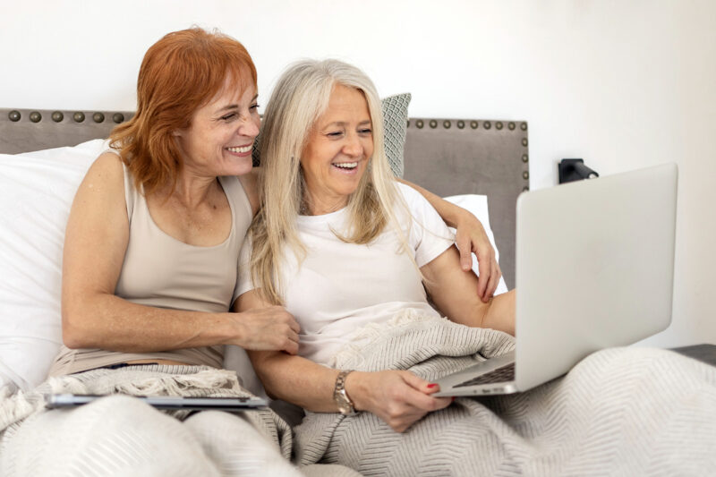 Two women in bed smiling and looking at a laptop